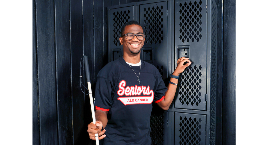 Christopher Morgan poses for a photo next to lockers while wearing his Alexander Senior Jersey and holding his cane.