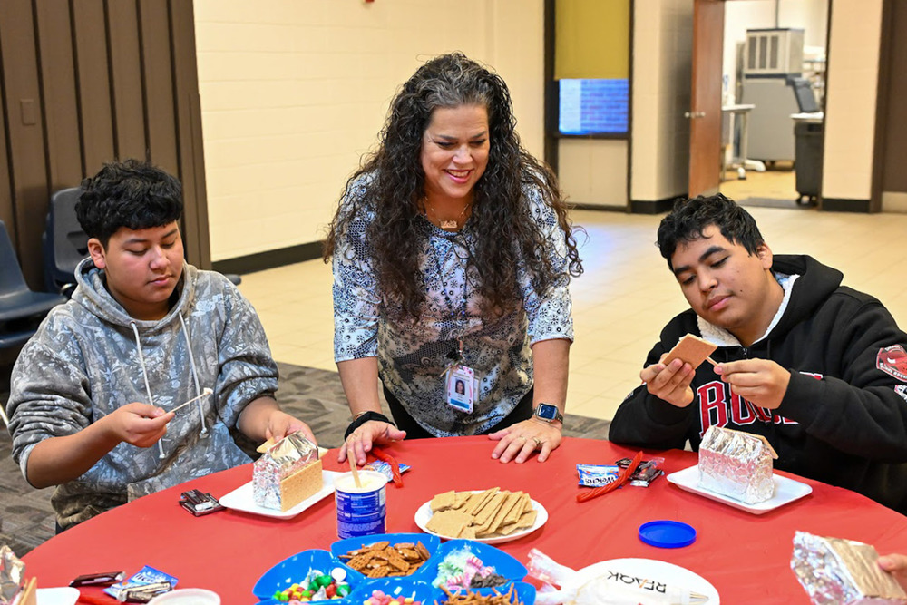 Dr. Araceli Hurley looks on as two Newcomer Center students work on their gingerbread houses.