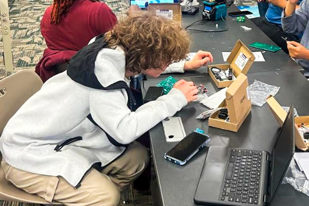A Lithia Springs High School student assembles electronic components for a DIY chatbot project, working with circuit boards and wiring at a lab table with a laptop nearby.