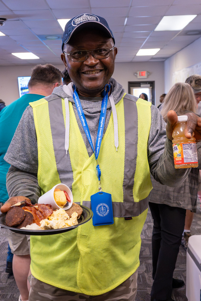 A DCSS bus driver wearing a yellow safety vest holds a plate of breakfast food.