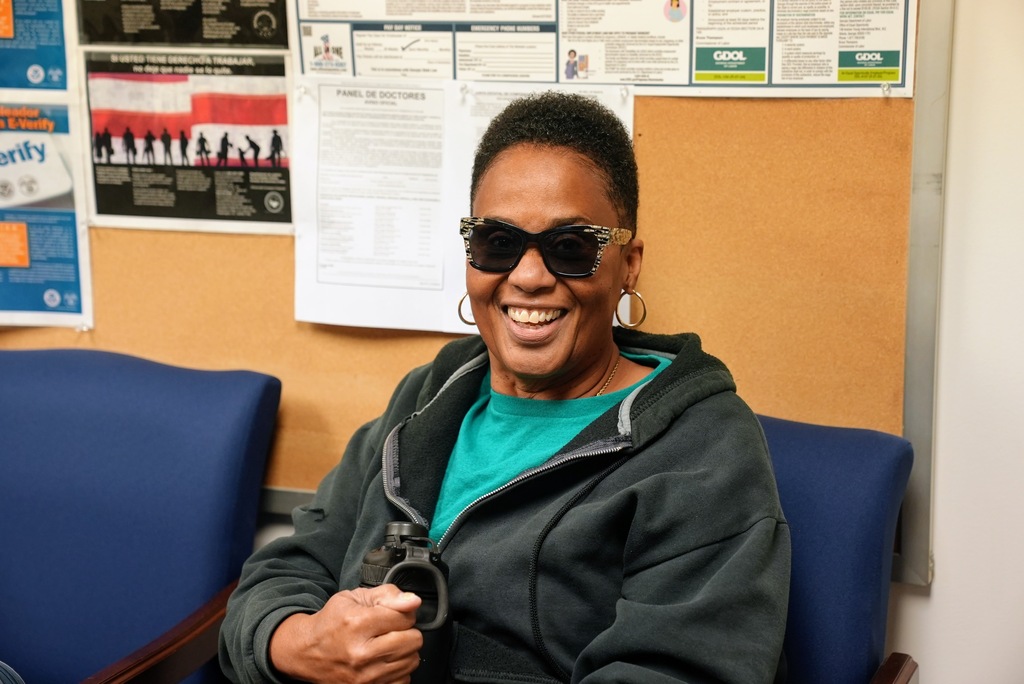 A bus driver wearingn a black jacket and sunglasses smiles.