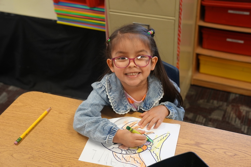 A student at Kinder Camp smiles while coloring a picture of a fish.