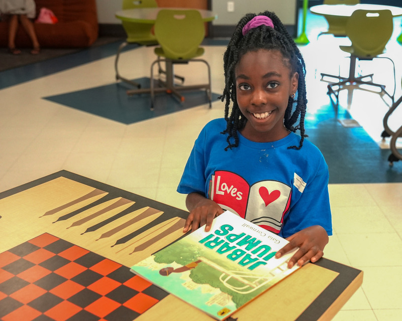 A Reading Ambassaord smiles while holding a book at a Summer Pop-Up event.