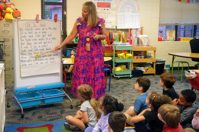 Teacher in a pink and purple floral dress stands in front of elementary students seated on classroom floor, gesturing while teaching a reading lesson displayed on chart paper showing a story titled "It Is Hot."