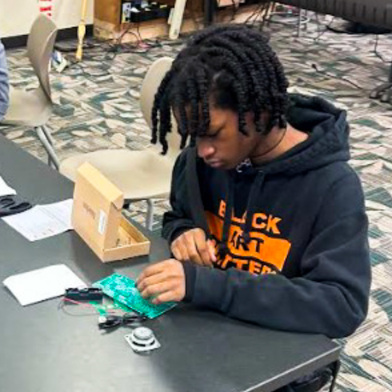 A Lithia Springs High School student wearing a "Black Art Matters" hoodie connects wiring to a green circuit board as part of a hands-on chatbot building project in Dr. Lindsey Napier's classroom.
