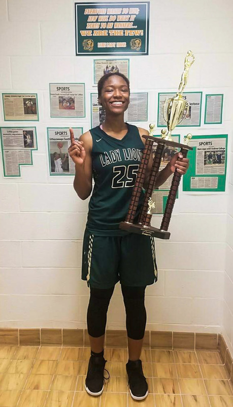 Lithia Springs' basketball legend Demeara Hinds holds a trophy while standing in front of a wall of newspaper clippings.