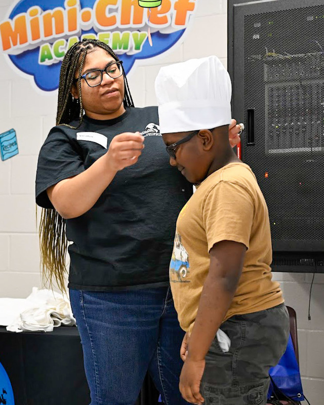 A member of the Rouse Foundation puts a chef's hat on a Sweetwater student during the Mini Chef Academy graduation.