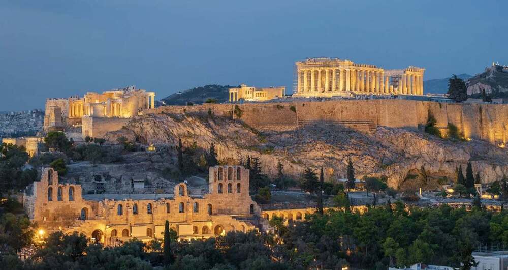 A wide view of the ancient Acropolis of Athens and the Parthenon, illuminated against a twilight sky.