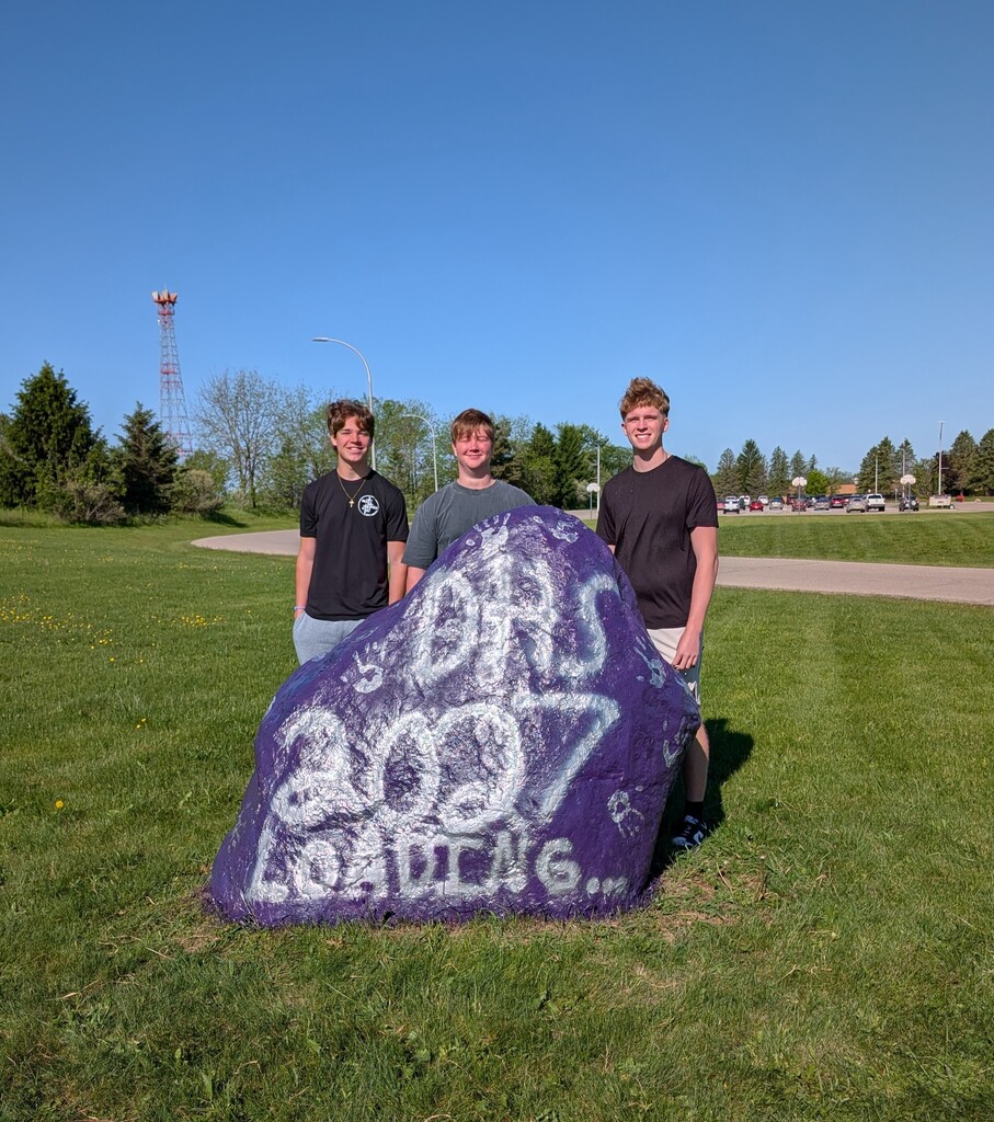 three high school juniors standing behind a painted boulder