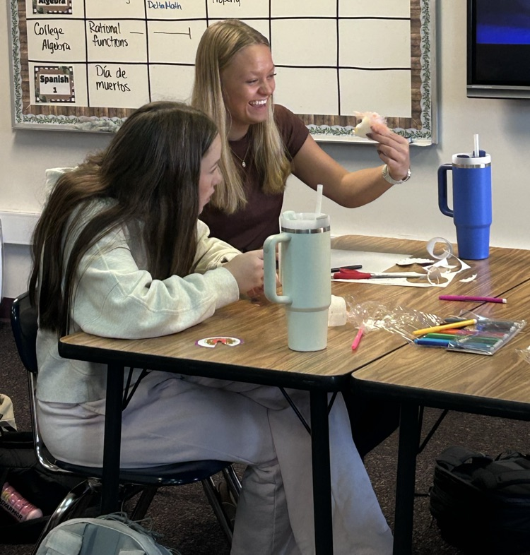 one student holds their sugar skull smiling and a second student looks across the table towards another student off camera 