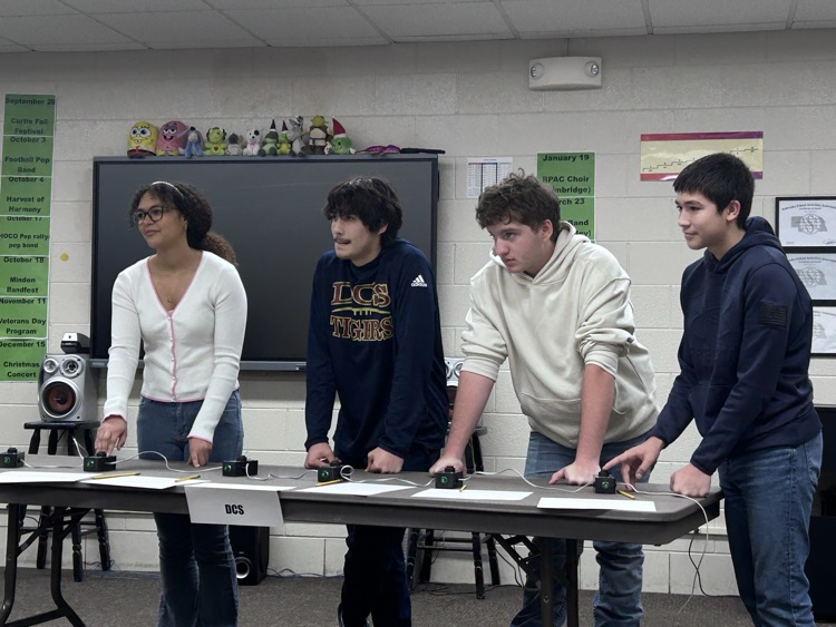 a team of 4 students stand behind a table listening to a question and prepare to buzz in