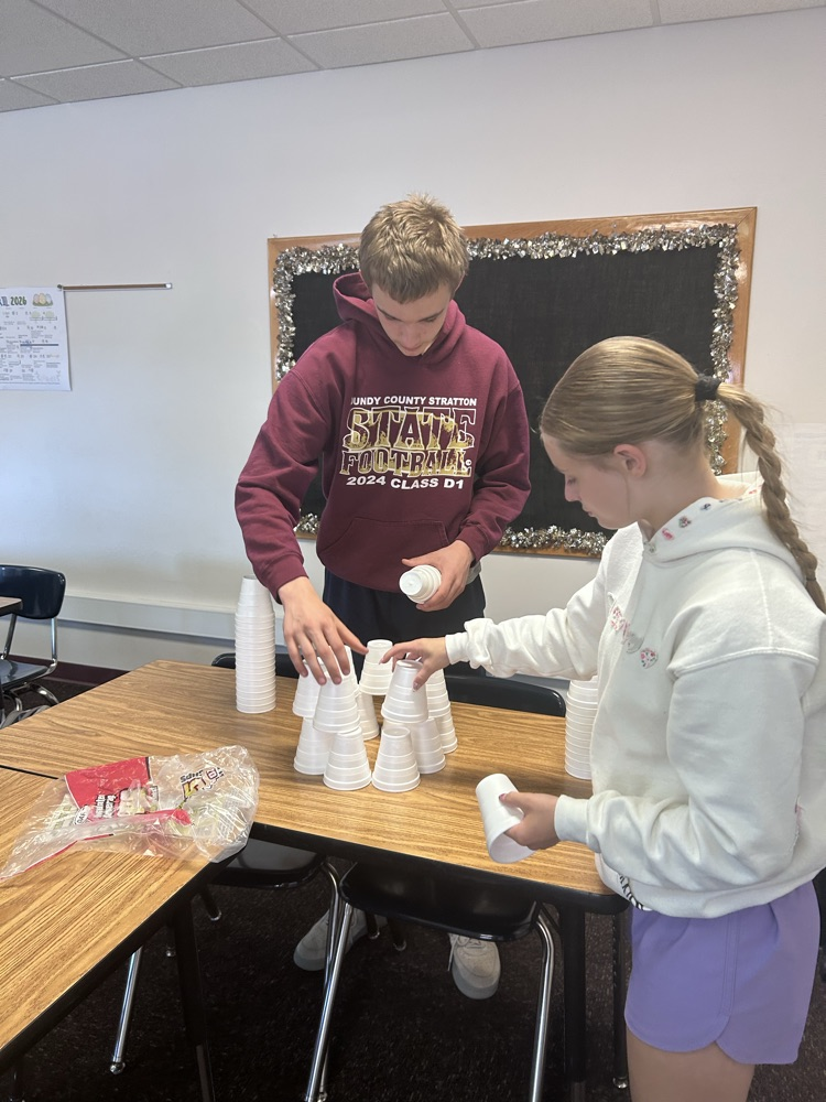 one male and one female student stand stacking styrofoam cups to form a round tower
