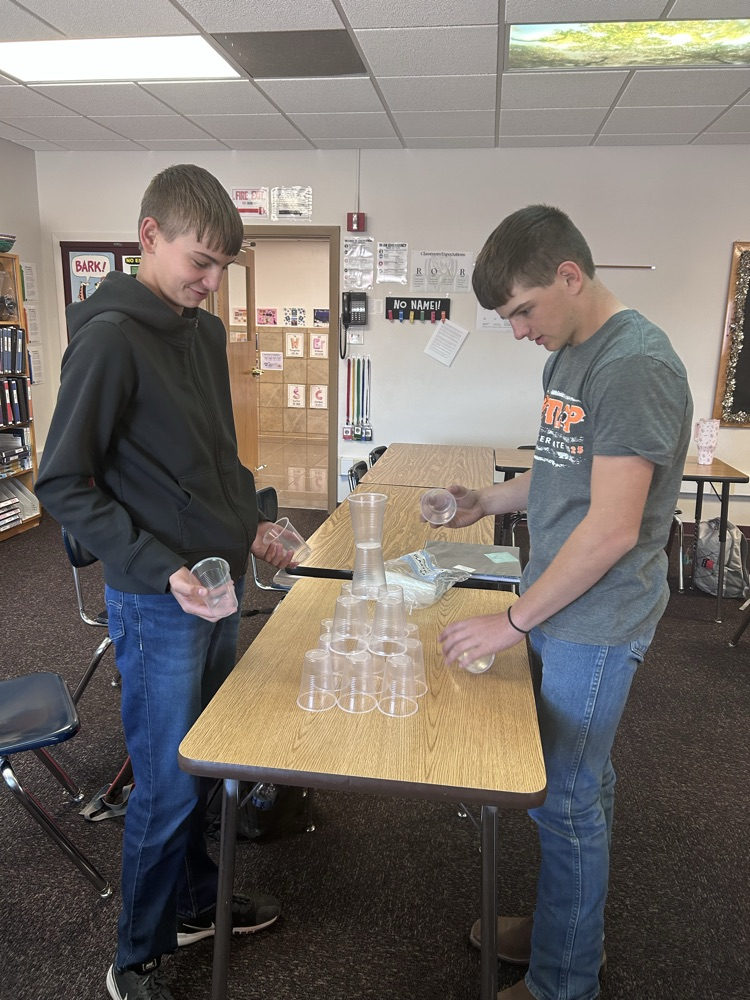 two male students stand building a tower with plastic cups on roof of a table