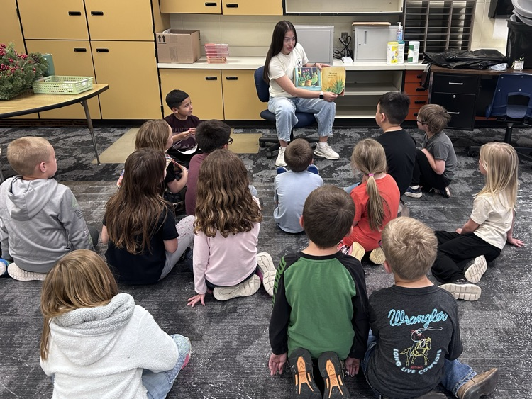 A female student sits in a chair with a book reading to the kindergarten class