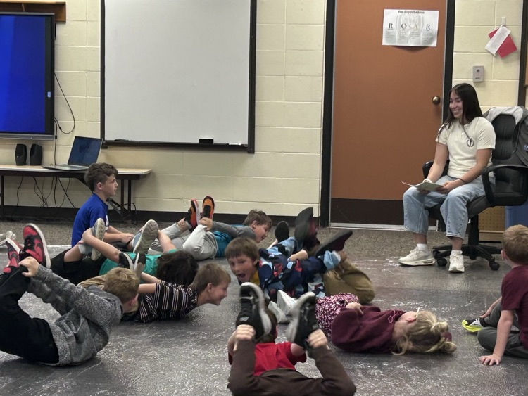 A female student sits in a chair laughing while the 1st grade student lay on the ground showing her how to do “the seal"