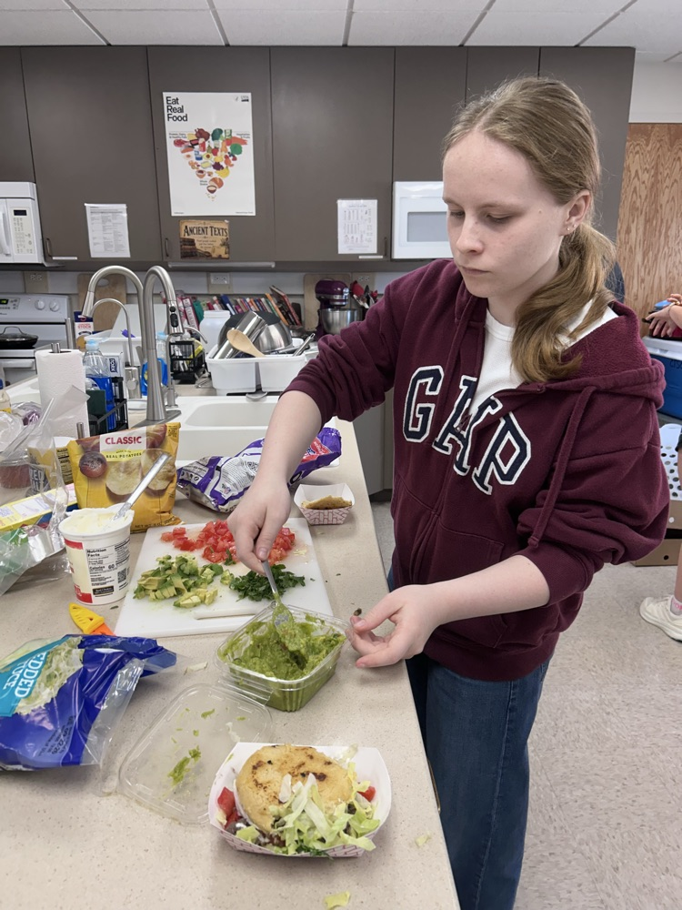 a female student wearing a maroon sweatshirt removes guacamole from a dish to prepare a Venezuelan style arepa
