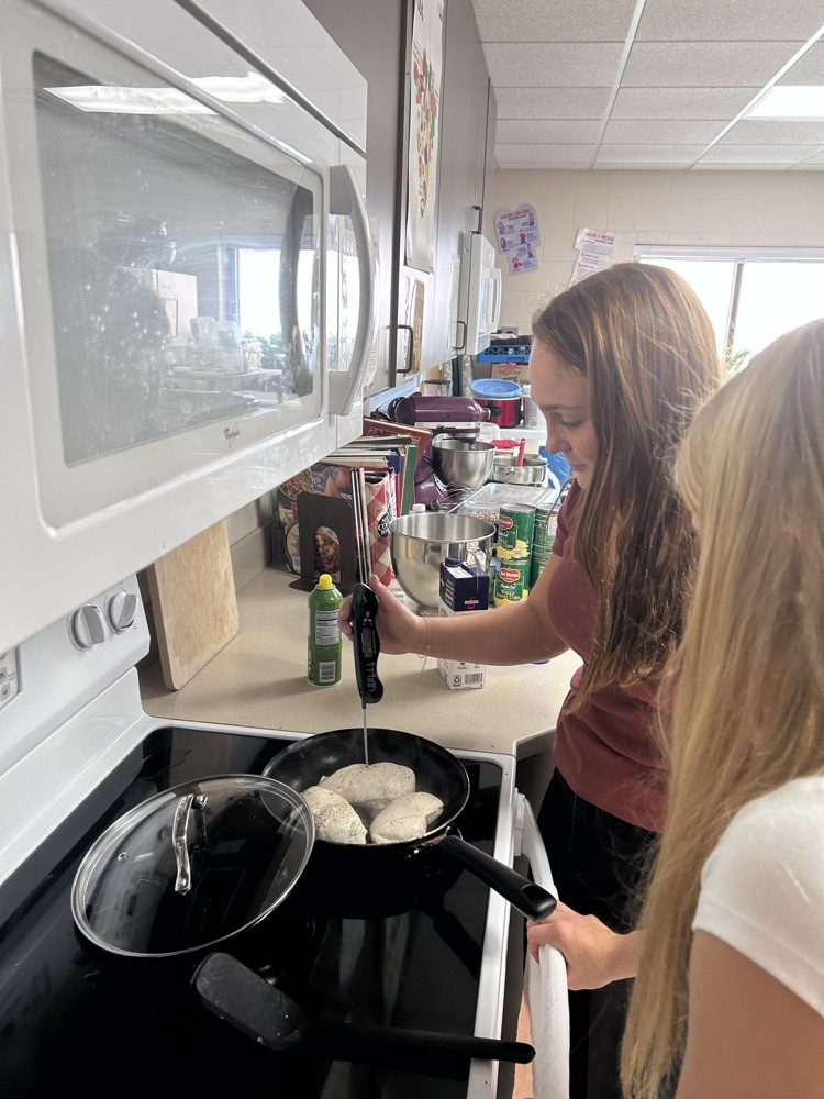 two female students are checking on the internal temperature of their chicken which is in a pan using a meat thermometer