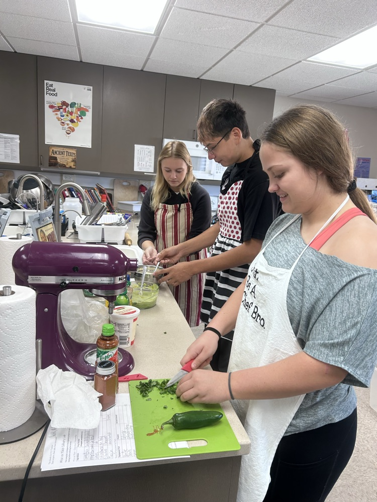 a female student wearing a white apron is chopping fresh cilantro on a green cutting board while a male and female student are in the background prepping their cheese and guacamole for their esquites recipe
