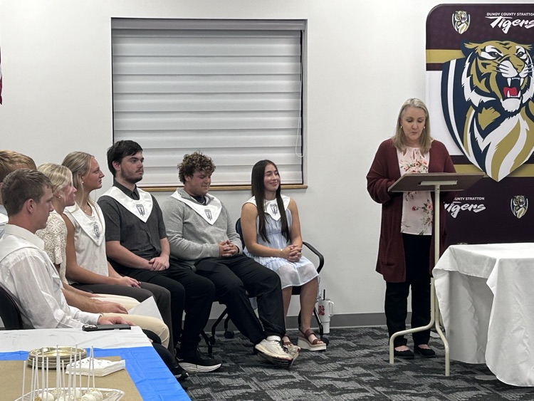 a teacher stand behind a podium speaking as 7 students sit listening