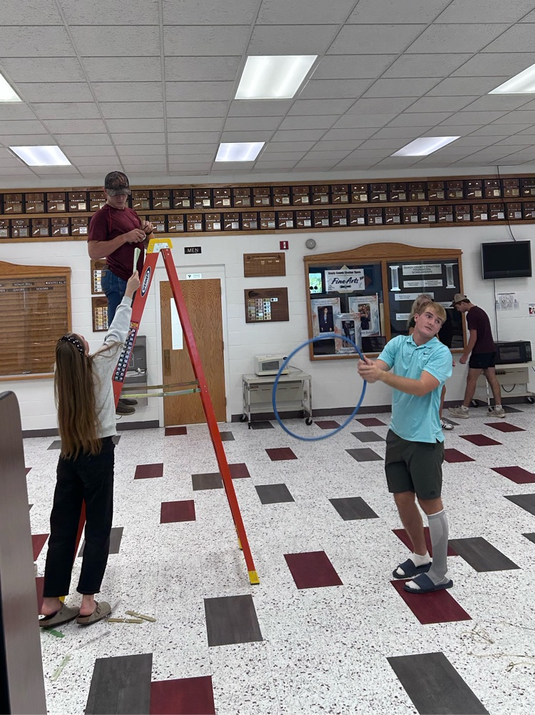 A male student is on a ladder with a female student handing him something and another male student holds a hula hoop