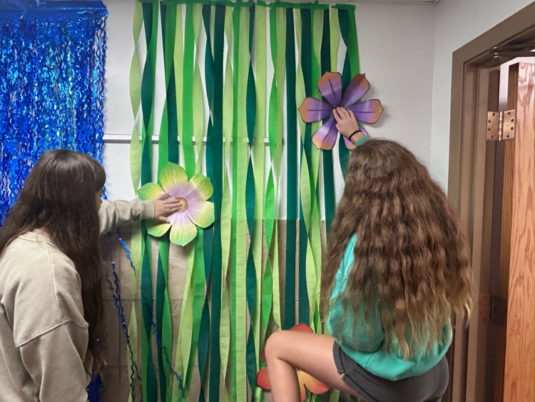 Two female students work to hang flowers on their wall of vines made with green crepe paper