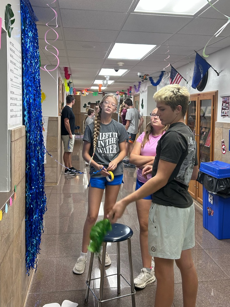 A male and two female students stand next to a wall discussing where to hang their tropical green leaves