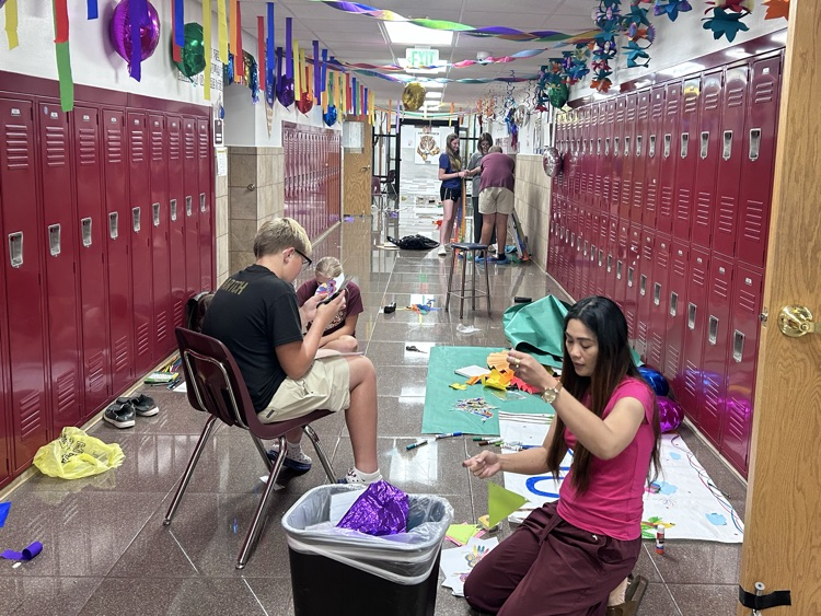 A teacher and a student sit in the foreground cutting out bright colored decorations. In the background are more students and colorful decorations hang from the ceiling