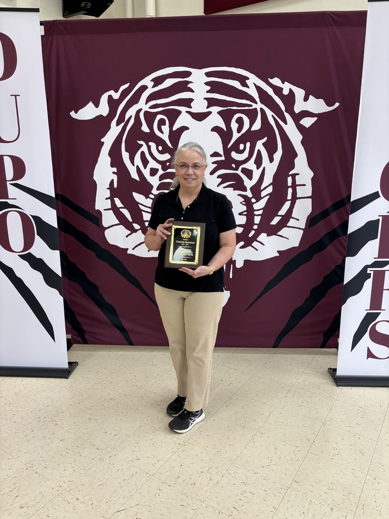 Person in black shirt and khaki pants holding plaque, standing in front of a maroon backdrop with a tiger logo.