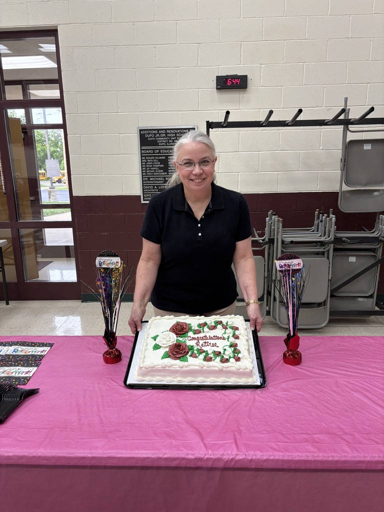 A woman stands behind a table with a pink tablecloth. In front of her is a cake with flowers on it.