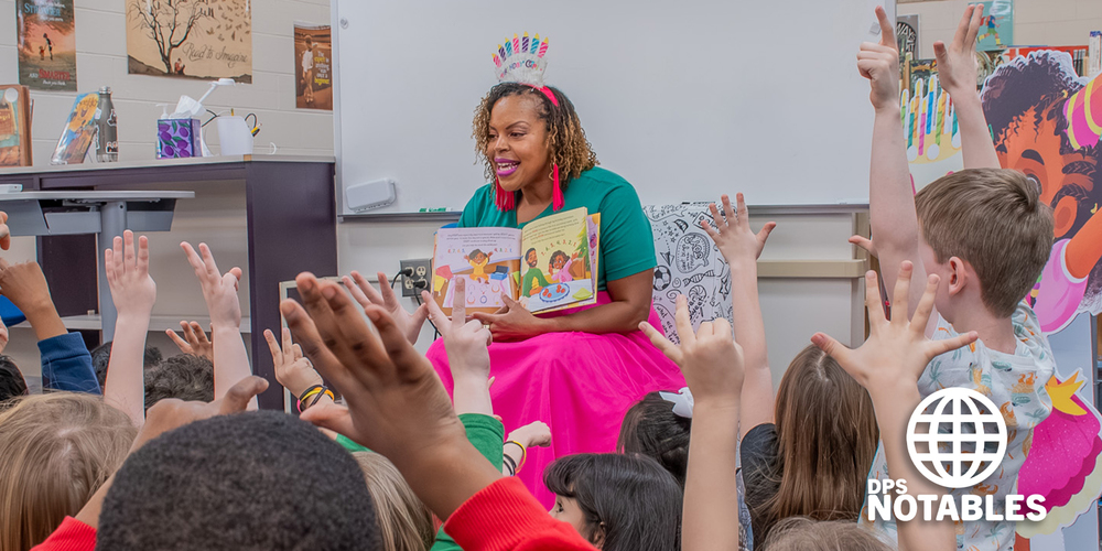A woman joyfully reads to children, holding an illustrated book. Kids eagerly raise their hands. The room is bright and lively, emphasizing learning.