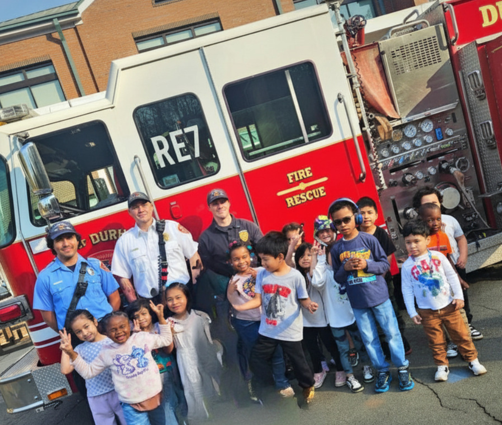 Group of firefighters and smiling children posing in front of a fire truck. The scene exudes joy and excitement, with kids showing playful gestures.