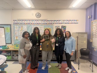 Five people stand in a classroom, smiling. One in the center holds a plaque and flowers. A colorful bulletin board is visible in the background.