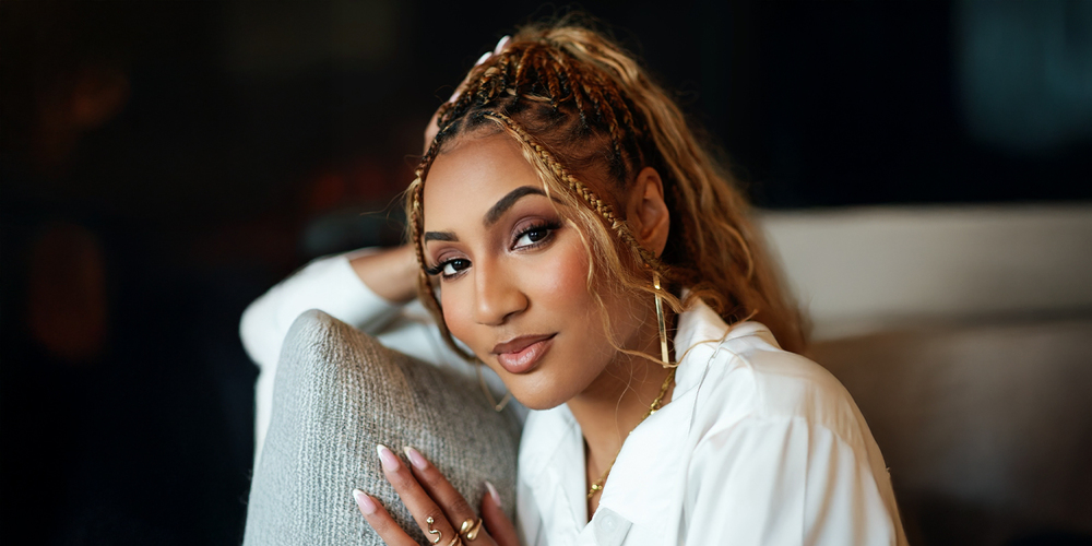 A woman with braided hair and a gentle smile leans on a gray cushion, wearing a white shirt. The setting is cozy, evoking a sense of relaxation and warmth.