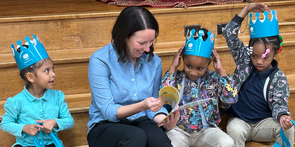 A teacher reads to three young children wearing blue paper crowns on wooden steps; two adjust their crowns as they listen, creating a warm, playful classroom scene.