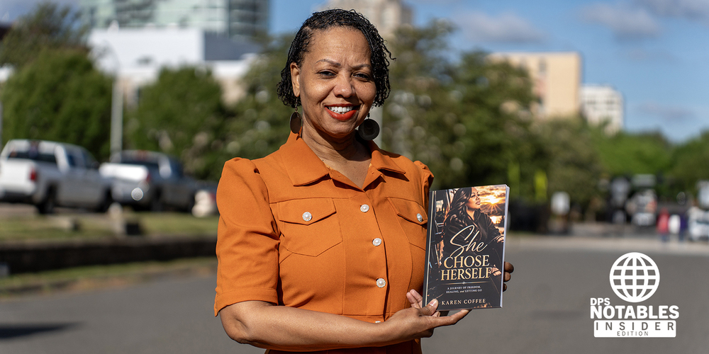 A woman in an orange dress smiles while holding a book titled "She Chose Herself." She's standing on a street with blurred buildings in the background.