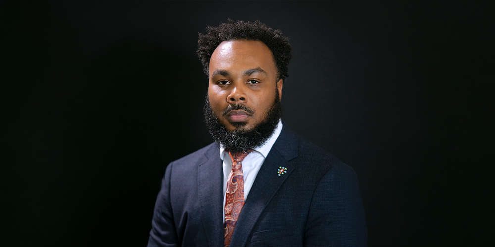 A man with a beard and short hair in a navy suit, white shirt, and patterned tie is posed against a dark background, conveying a professional tone.