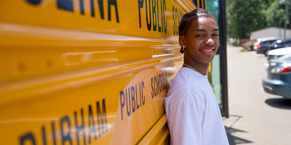 Smiling teenager in a white shirt leans against a yellow school bus with visible text "Public Schools." Bright, sunny day, relaxed atmosphere.