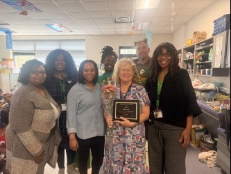 A group of seven people stands in a classroom, smiling. One person in front holds a plaque and a flower bouquet, signifying an award celebration.