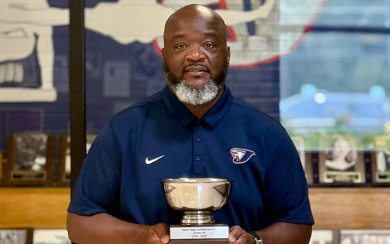 A person holding a trophy stands in front of display shelves with framed photos. They wear a navy shirt with a logo, exuding pride and accomplishment.