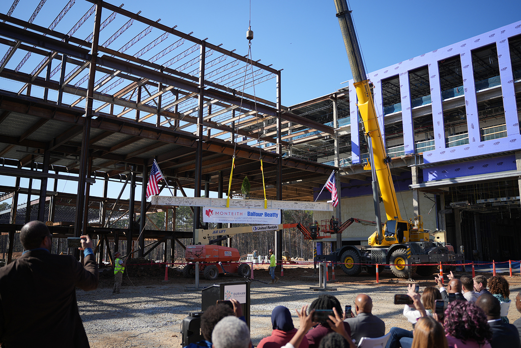 A construction site with a large steel beam being lifted by a yellow crane. American flags are displayed, and a group of people watches, capturing photos. The atmosphere is focused and anticipatory under a clear blue sky.