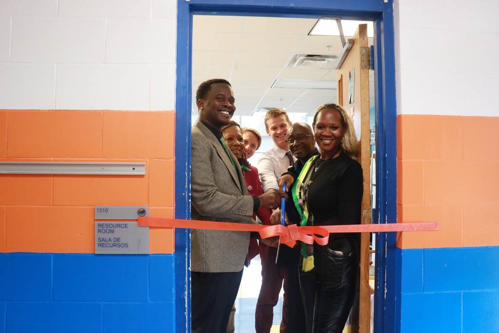 A group of people smiles while cutting a red ribbon across a doorway labeled "Resource Room" in a brightly colored hallway, signifying a grand opening.