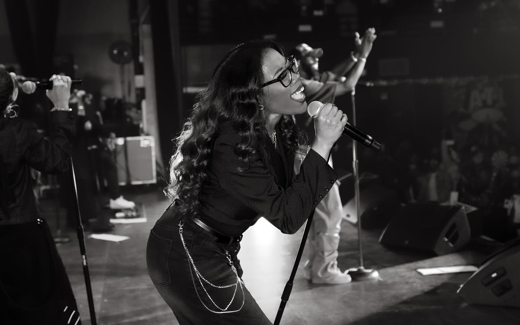 Black and white image of a woman on stage, singing passionately into a microphone. She has long wavy hair, wears glasses, and has a joyful expression.