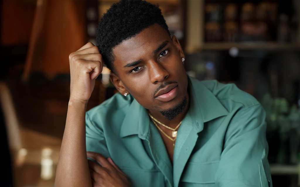 A man in a green shirt sits thoughtfully at a wooden table, with his elbow resting and hand on his temple. Soft focus background, warm and contemplative tone.