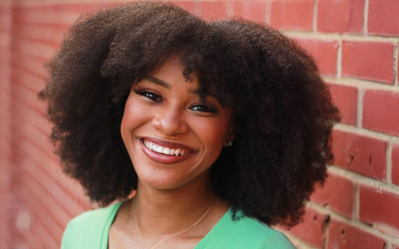 Smiling woman with curly hair wearing a green top stands in front of a red brick wall. The tone is joyful and warm.