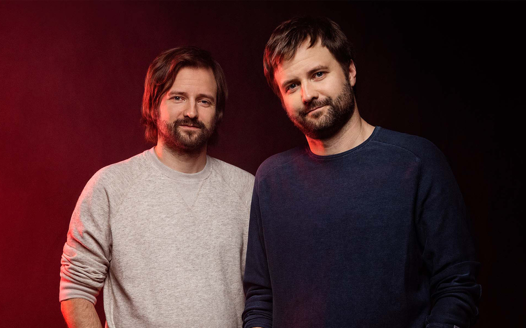 Two men with beards stand closely against a dark gradient background with red lighting. One wears a gray sweater, the other a navy shirt, both smiling subtly.