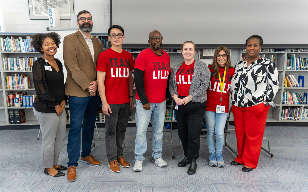 Seven adults stand in a library. Four wear red "Team Lilly" shirts, conveying teamwork and unity. Bookshelves are visible, suggesting a scholarly setting.
