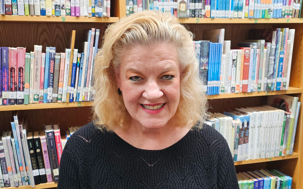 A woman with curly blond hair smiles warmly in a library setting, surrounded by colorful books on wooden shelves, conveying a welcoming tone.