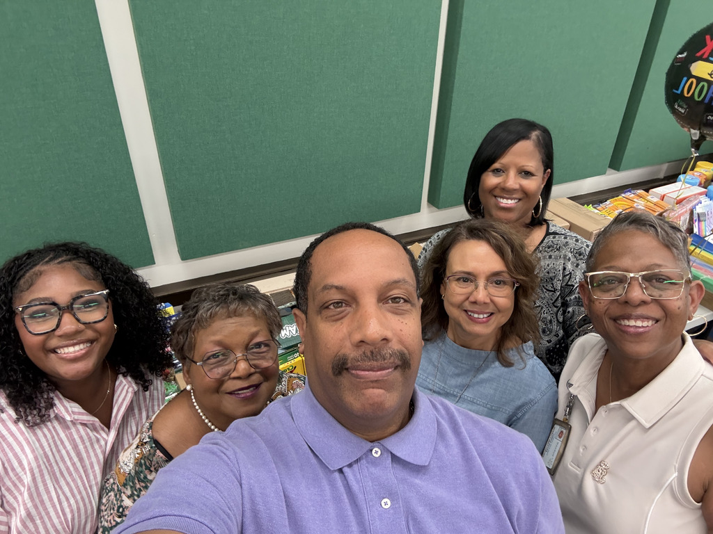 Five school staff members pose together for a selfie in front of tables filled with donated school supplies.
