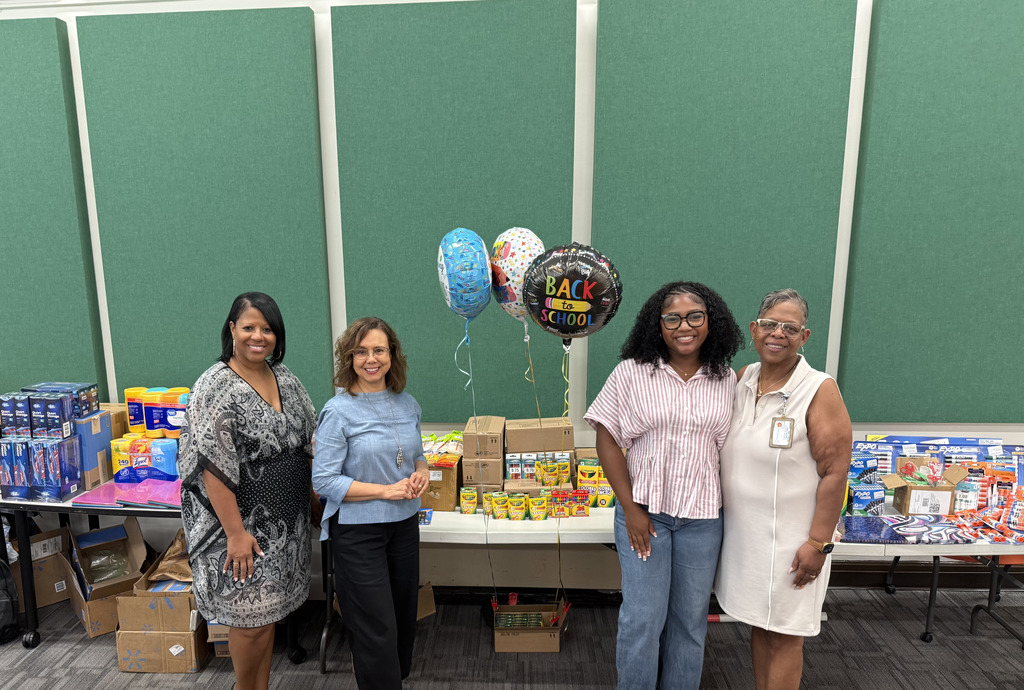Four school staff members pose together in front of tables displaying donated school supplies and “Back to School” balloons.