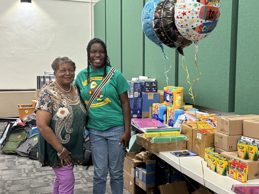Adults stand together beside tables filled with donated school supplies and backpacks, with colorful “Back to School” balloons overhead.